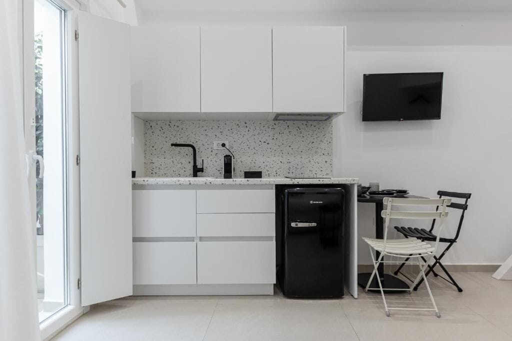 A compact kitchen with white cabinetry, a built-in dishwasher, a small black refrigerator, and a wall-mounted TV.