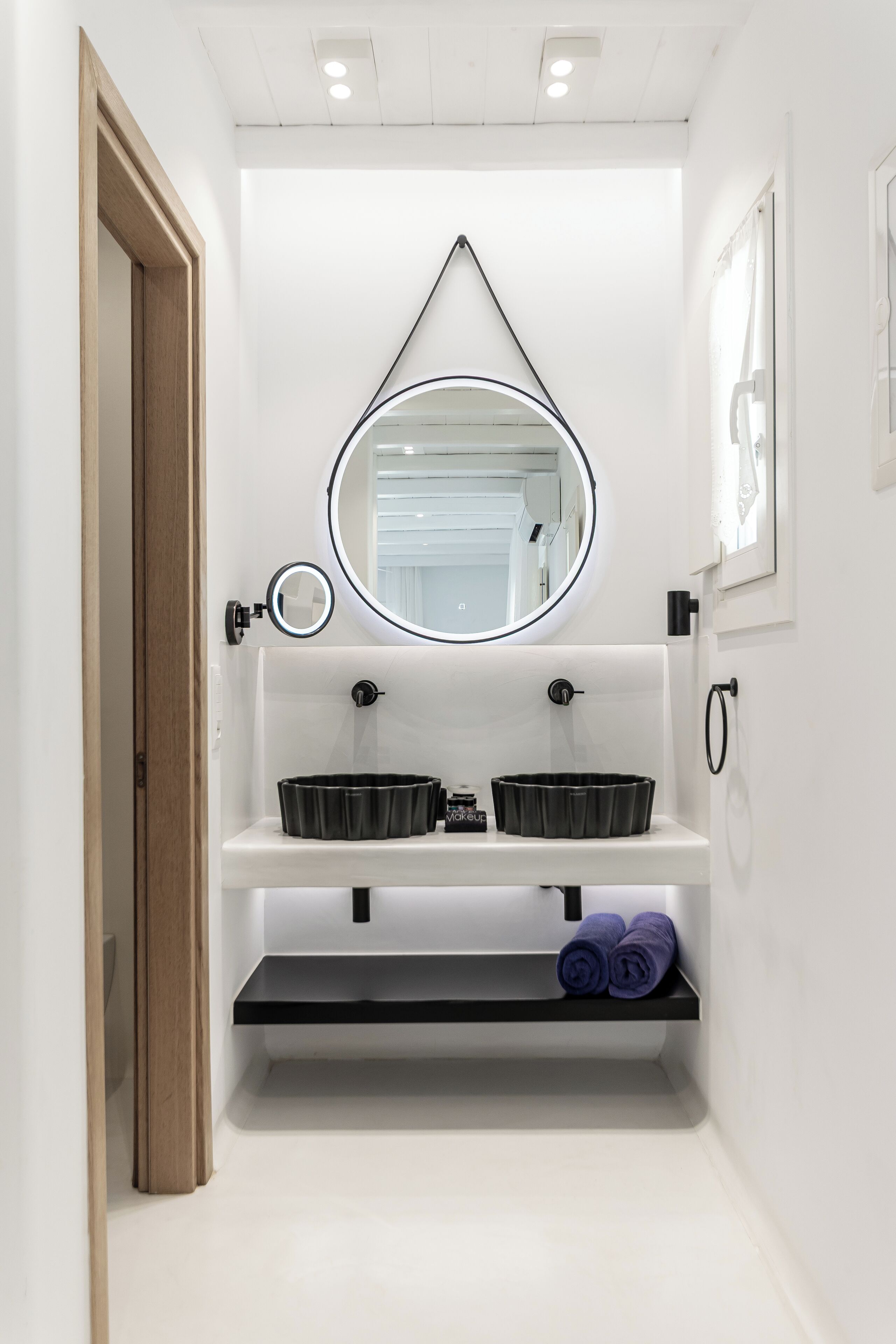 A modern bathroom with a white vanity, two black sinks, a round mirror, and a wooden door.