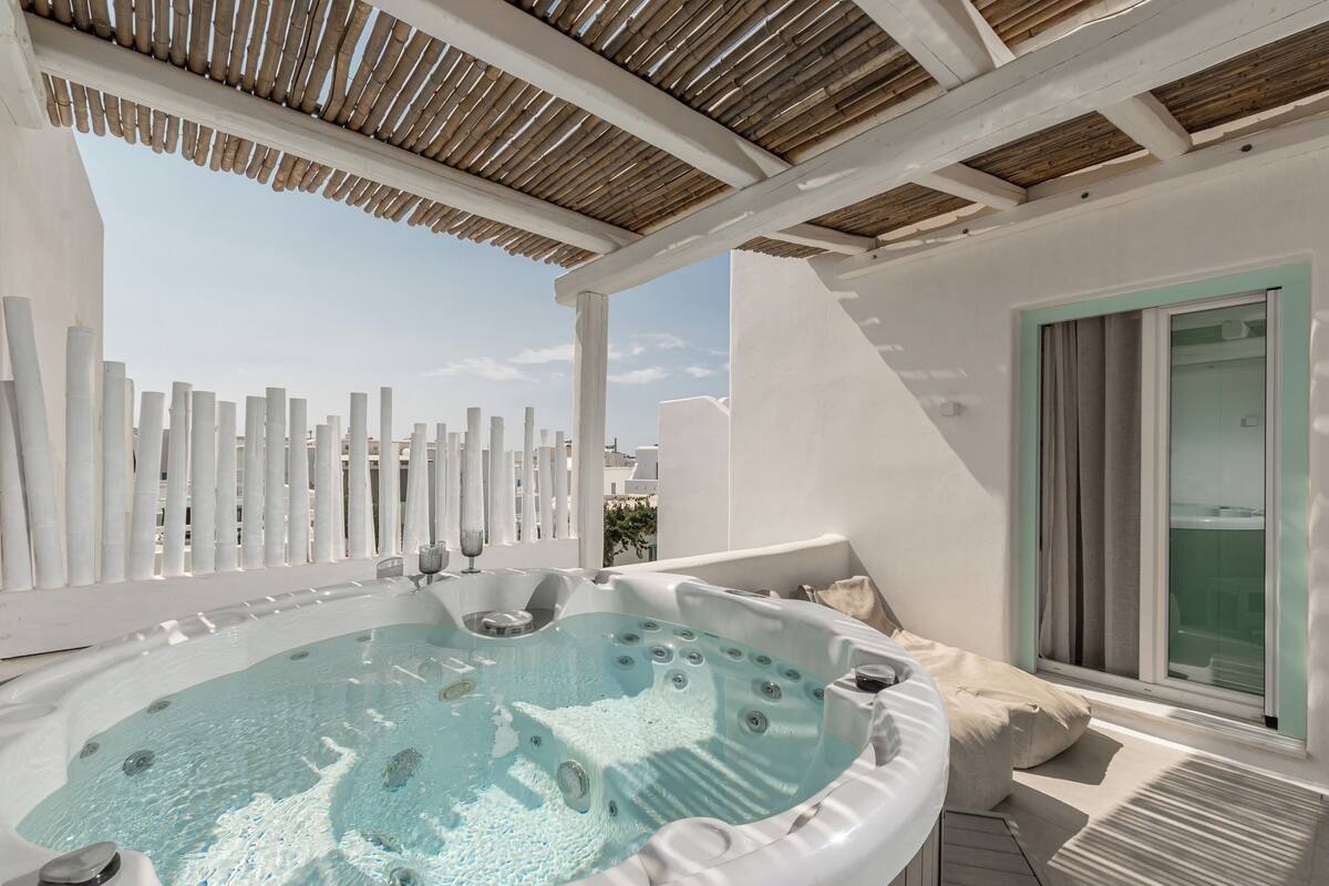 Ein Whirlpool unter einer Holzpergola mit einem weißen Zaun, einem Fenster mit Vorhängen und Blick auf Gebäude.