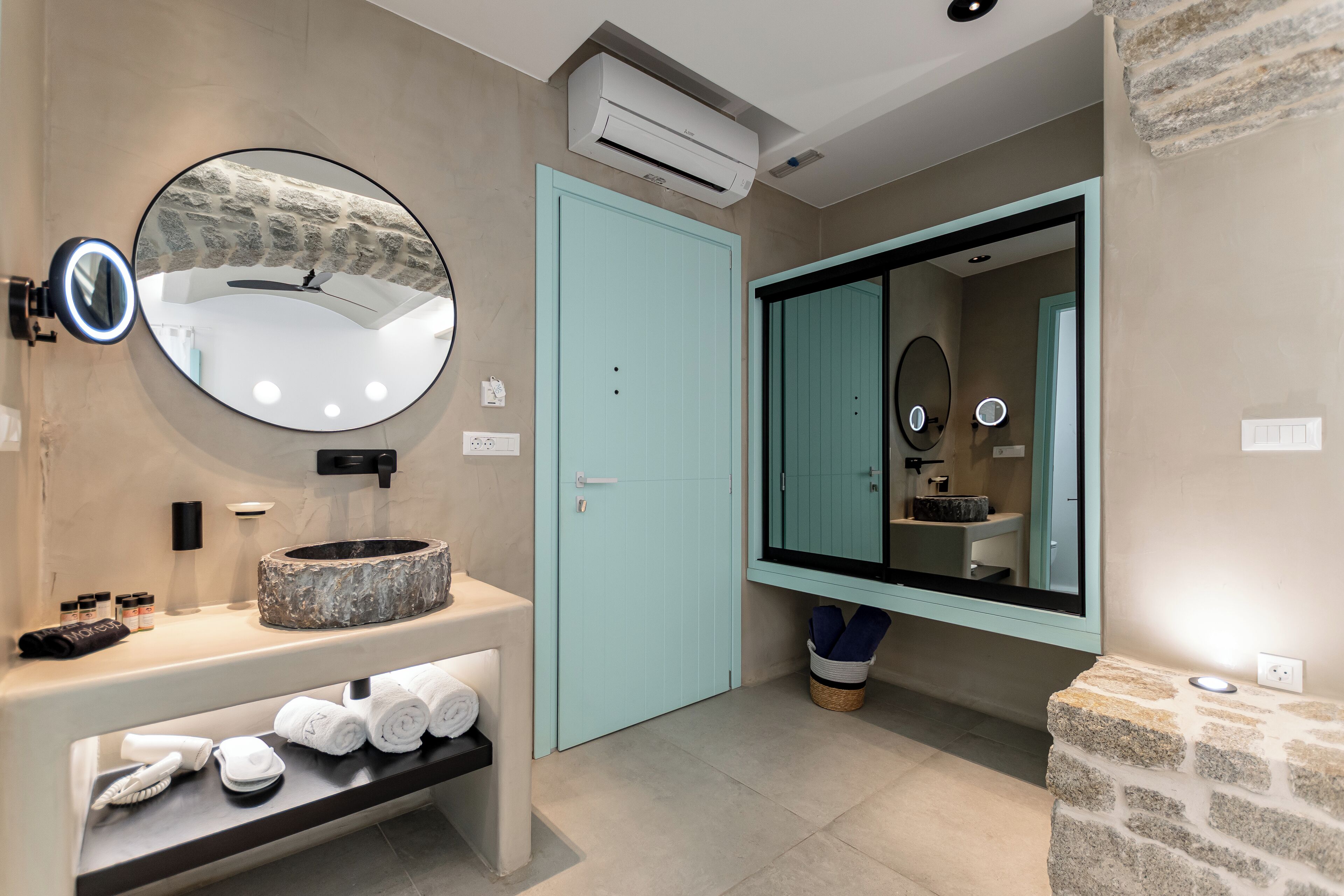 Une salle de bain moderne avec un lavabo en pierre, un grand miroir rond doté de lumières, une porte de couleur turquoise et un grand miroir au-dessus du lavabo.