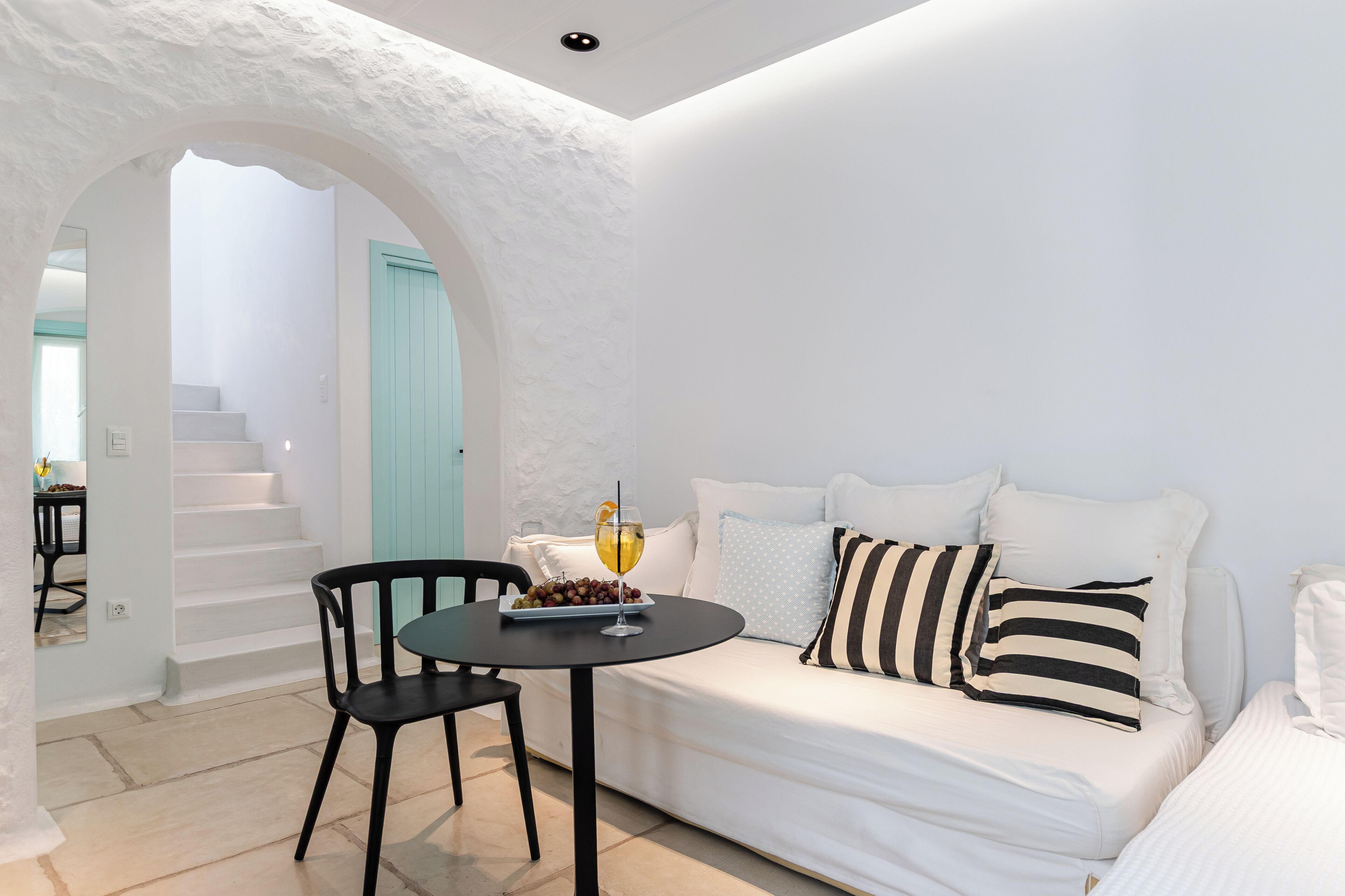 A white-walled room with a white sofa, black and white striped pillows, a table with a glass of drink, and a chair.