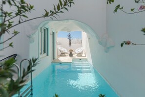 An indoor swimming pool with clear blue water, a glass archway, and two white lounge chairs with a small table in the background.