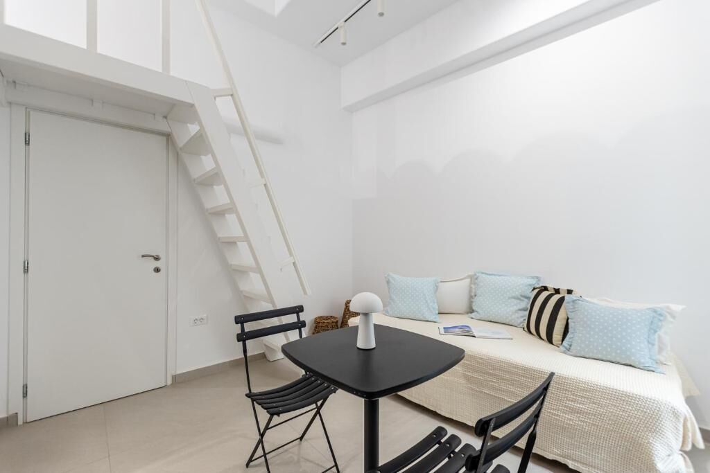 A compact room with a bed, a small table, and two chairs. There is a white door and a white ladder leaning against the wall.