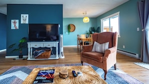 Un salon avec un téléviseur, un fauteuil marron, une table à manger avec des chaises et une grande fenêtre.