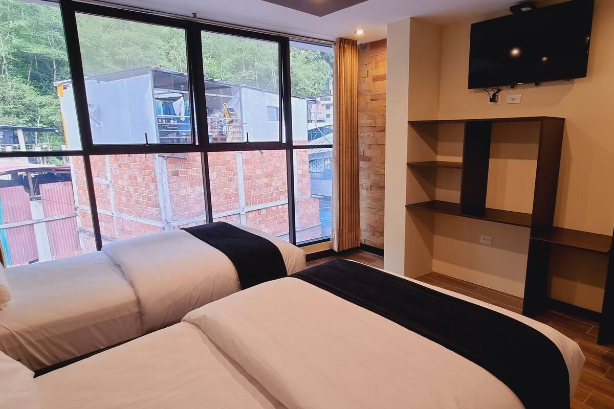 A hotel room with two beds, a TV mounted on the wall, and a view of a brick building outside.
