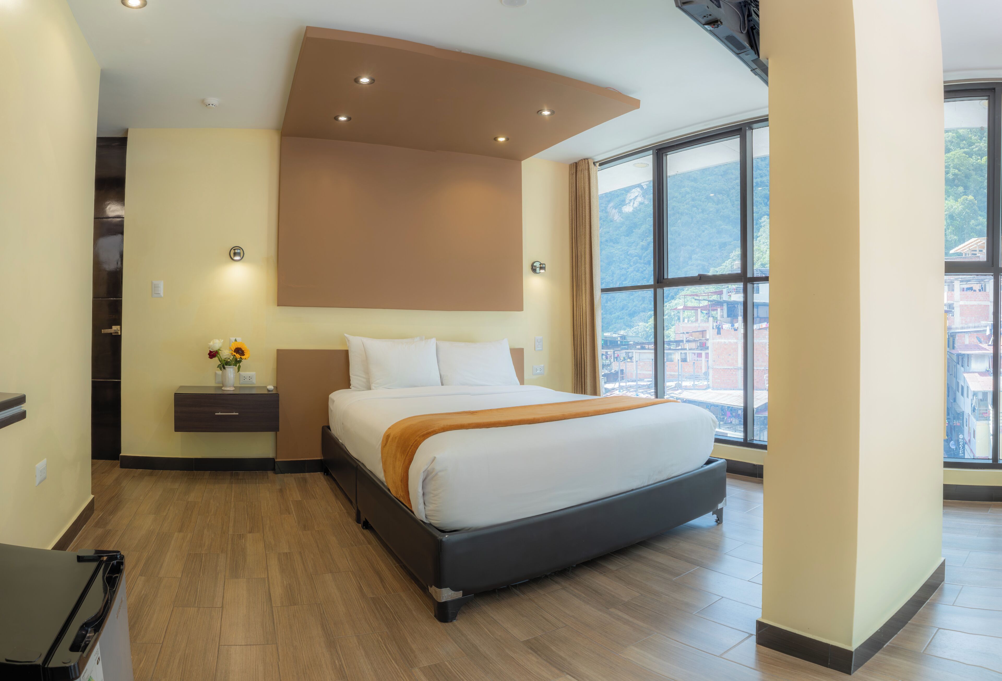 A modern hotel room with a large bed, a bedside table, a nightstand, a window with a city view, and a wooden floor.