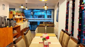 Un comedor con una mesa larga, sillas e iluminación colgante. Se observa una zona de buffet con estantes y vistas al exterior.