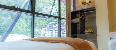 Una habitación de hotel con un ventanal grande, una cama con una manta marrón, un estante con fruteros y un jarrón con flores.