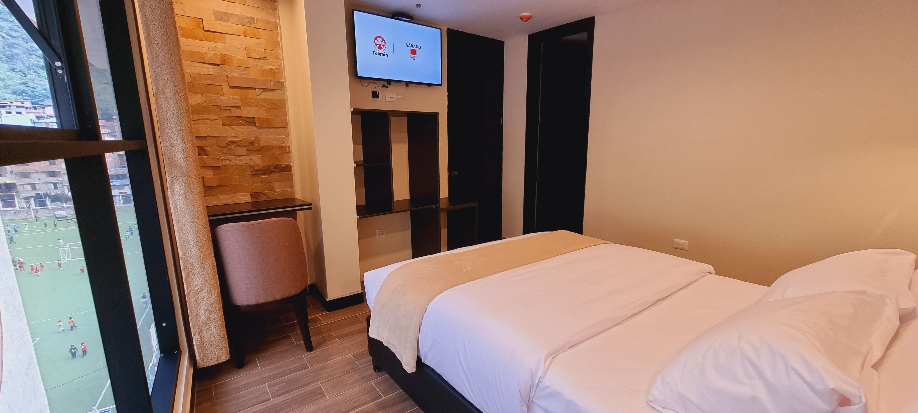Quarto de hotel com cama, escrivaninha, cadeira, TV fixada na parede e janela com vista para um campo de futebol.