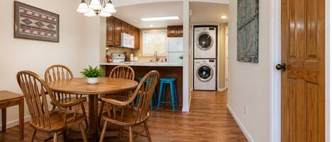 A kitchen with wooden cabinets, a dining table with chairs, a hanging light fixture, and a washing machine in the background.