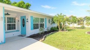 A single-story house with a front porch, a blue door, and a small garden area with palm trees. - Pine Island Beach House!  *Gulf Access, Heated Pool, Hot Tub & Boat Dock* (St. James City)