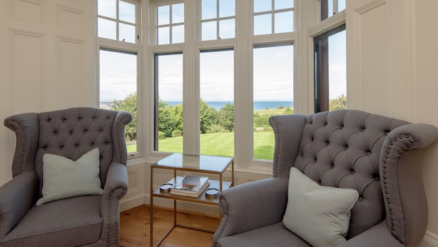 A room with two grey upholstered armchairs, a glass coffee table, and large windows offering a view of the sea. - A unique, spectacular detached home in a much sought-after area with unbeatable panoramic sea views. (North Berwick)