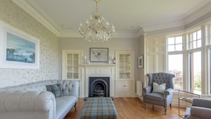 A spacious living room with a fireplace, a chandelier, and large windows. - A unique, spectacular detached home in a much sought-after area with unbeatable panoramic sea views. (North Berwick)