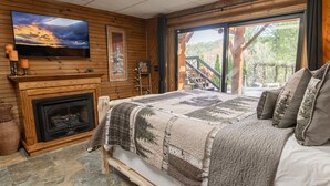Ein Schlafzimmer mit einem großen Bett, einem Kamin, einem an der Wand befestigten Fernseher und Blick auf eine Holzterrasse und Bäume.