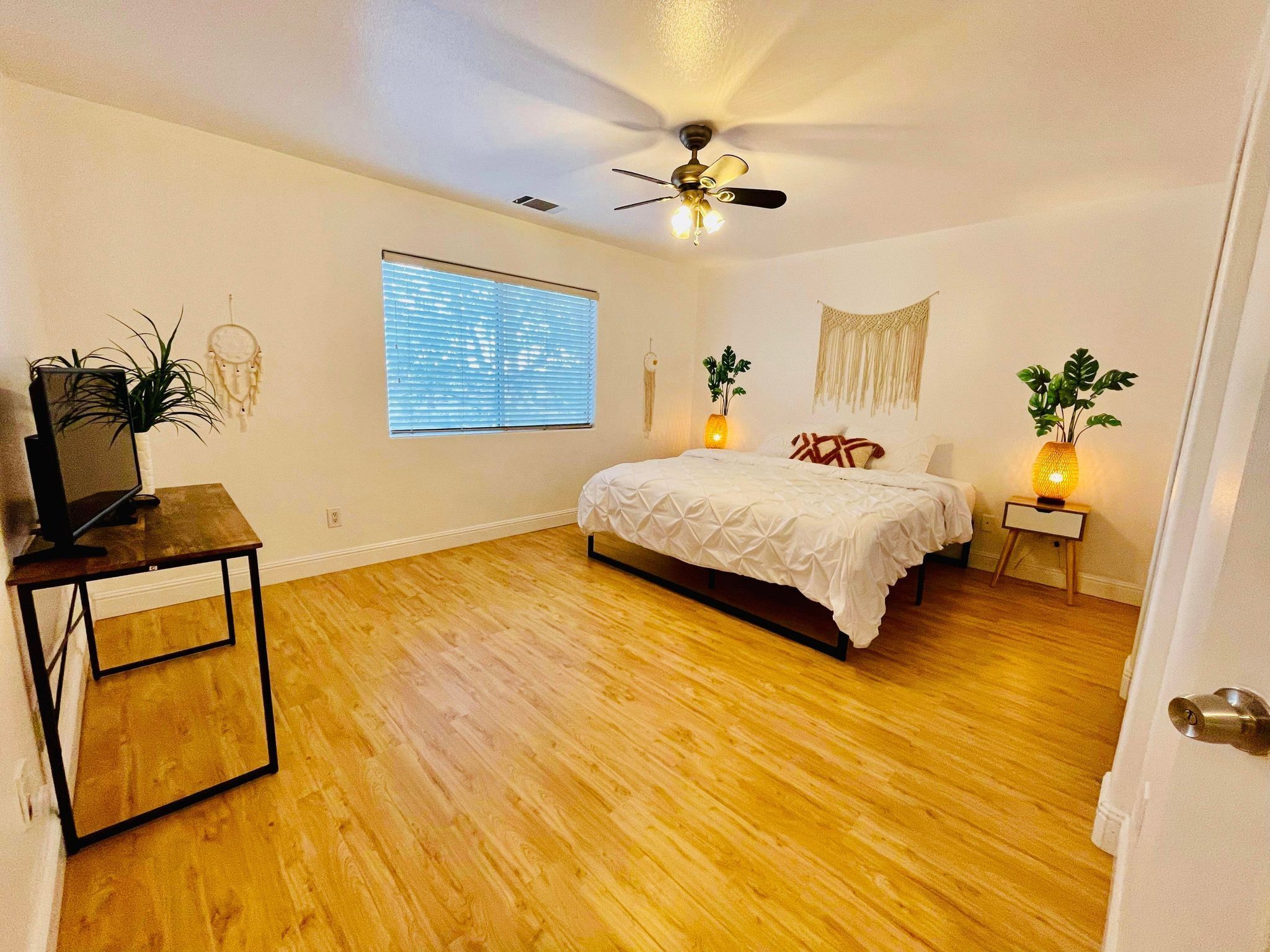 Une chambre à coucher avec un lit, un ventilateur de plafond, une fenêtre avec des stores, une table de chevet avec une lampe et une petite plante.
