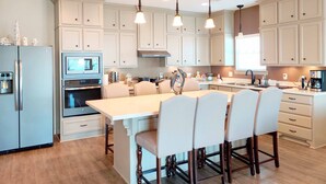 A modern kitchen with white cabinets, a central island with chairs, a refrigerator, oven, and a coffee maker.