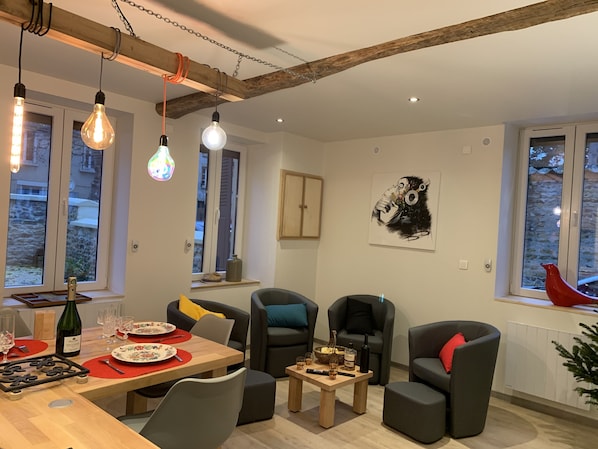 A modern living room with a dining area, featuring a wooden table set for a meal, comfortable seating, and large windows with a view.