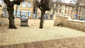 A wooden deck with a gravel area, trees, a metal gate, and stone buildings in the background. - Gîte Le BISTANCLAC in the heart of the town of CHARLIEU. (Charlieu)