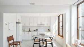 A modern kitchen with white cabinets, a dining table with four chairs, a wooden chair, and a sofa with cushions. - Le Nomade MTL (Montreal)