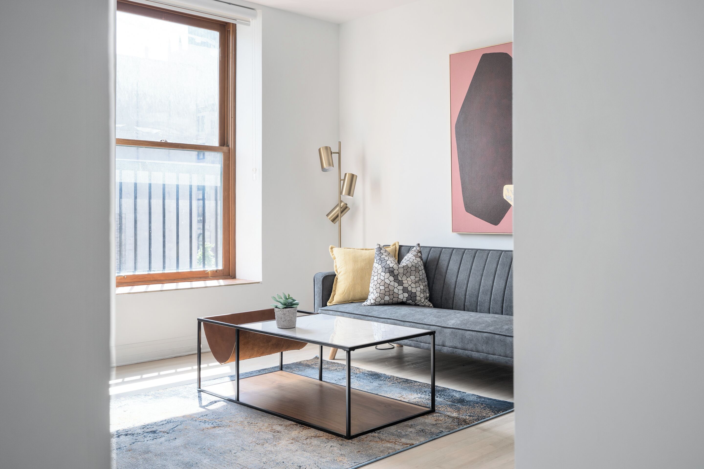 Un salón moderno con una mesa de centro de vidrio, un sofá gris con cojines estampados, un ventanal grande y una obra de arte minimalista en la pared.