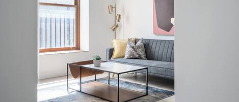 Un salón moderno con una mesa de centro de vidrio, un sofá gris con cojines estampados, un ventanal grande y una obra de arte minimalista en la pared.