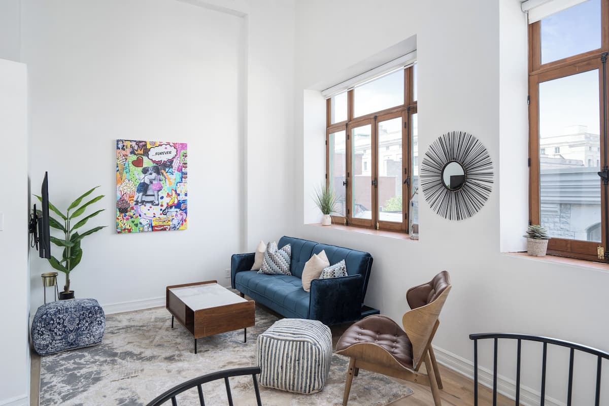 A modern living room with a blue sofa, a coffee table, a striped ottoman, a brown chair, and a black metal chair. There is a colorful artwork on the wall, a potted plant, and a sunburst mirror on the wall.