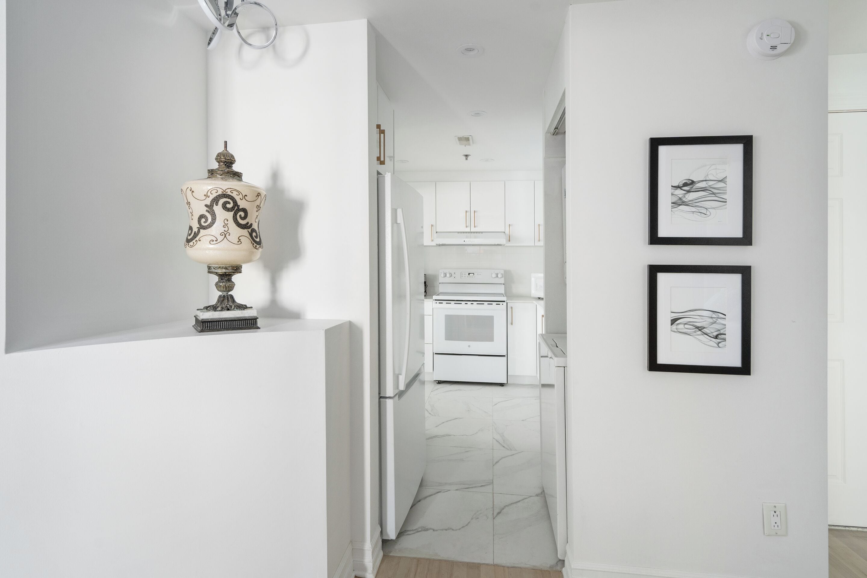 A narrow hallway with white walls, a marble floor, a white refrigerator, a white oven, and two framed abstract artworks.