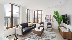 A modern living room with a sofa, chairs, a coffee table, and a TV. Large windows provide natural light and a view of the city. - Le Nomade MTL (Montreal)