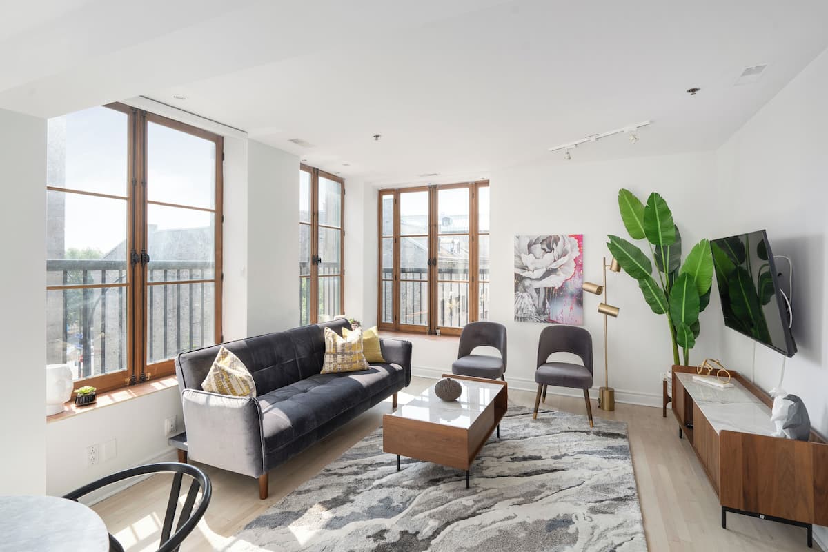 A modern living room with a sofa, chairs, a coffee table, and a TV. Large windows provide natural light and a view of the city.