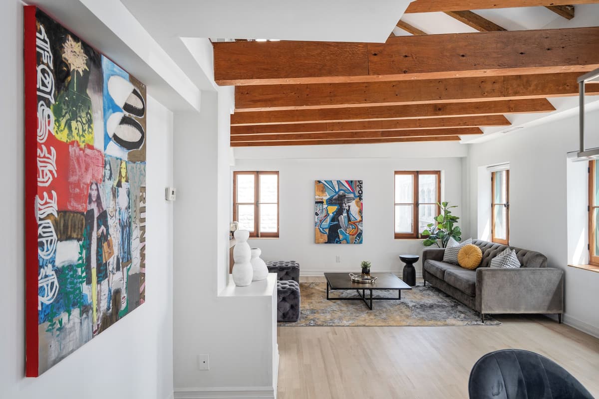 A spacious living room with a high ceiling, wooden beams, a grey sofa, a coffee table, and two large paintings on the walls.