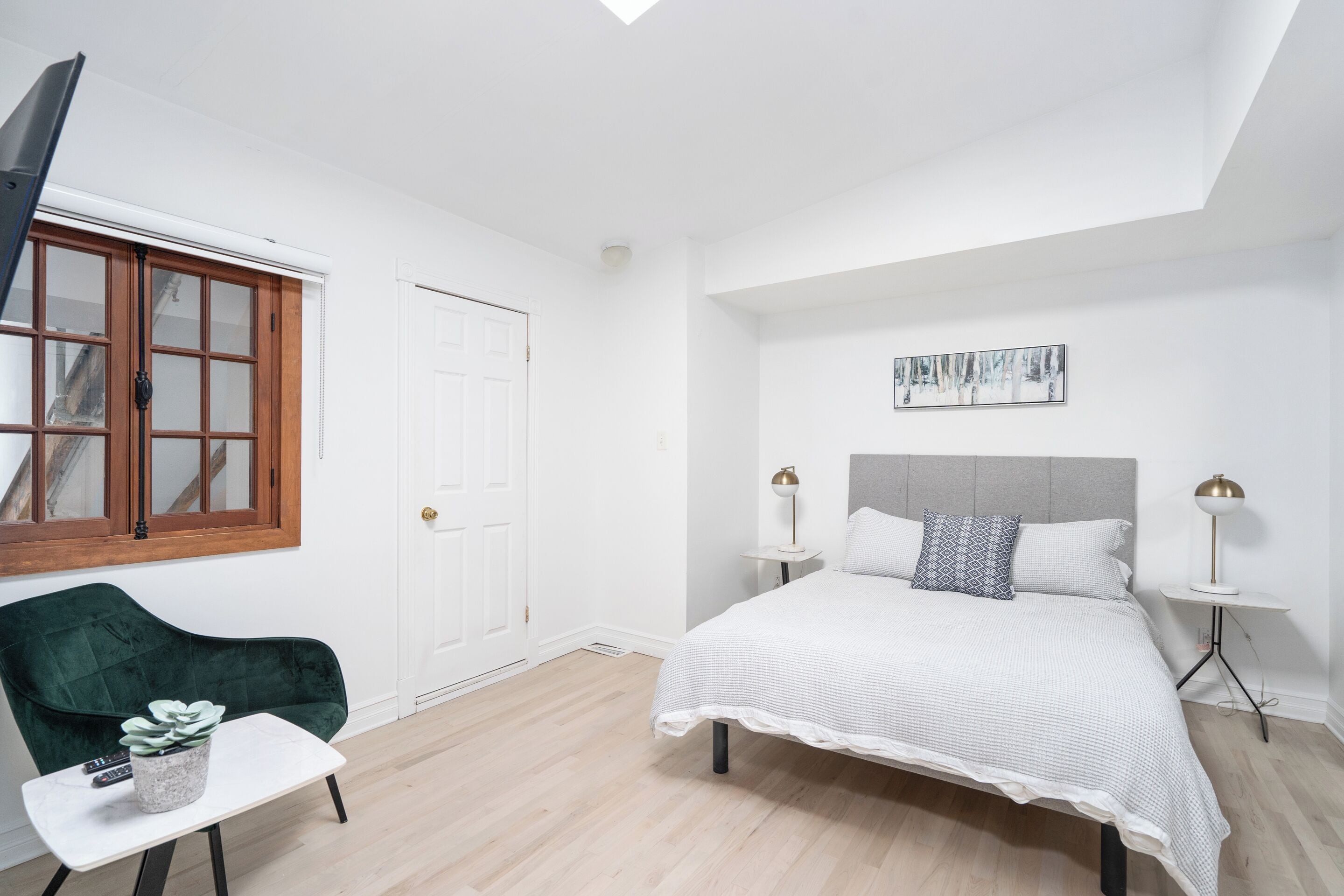 A modern bedroom with a bed, a green chair, a small table with a plant, and a window with wooden frames.