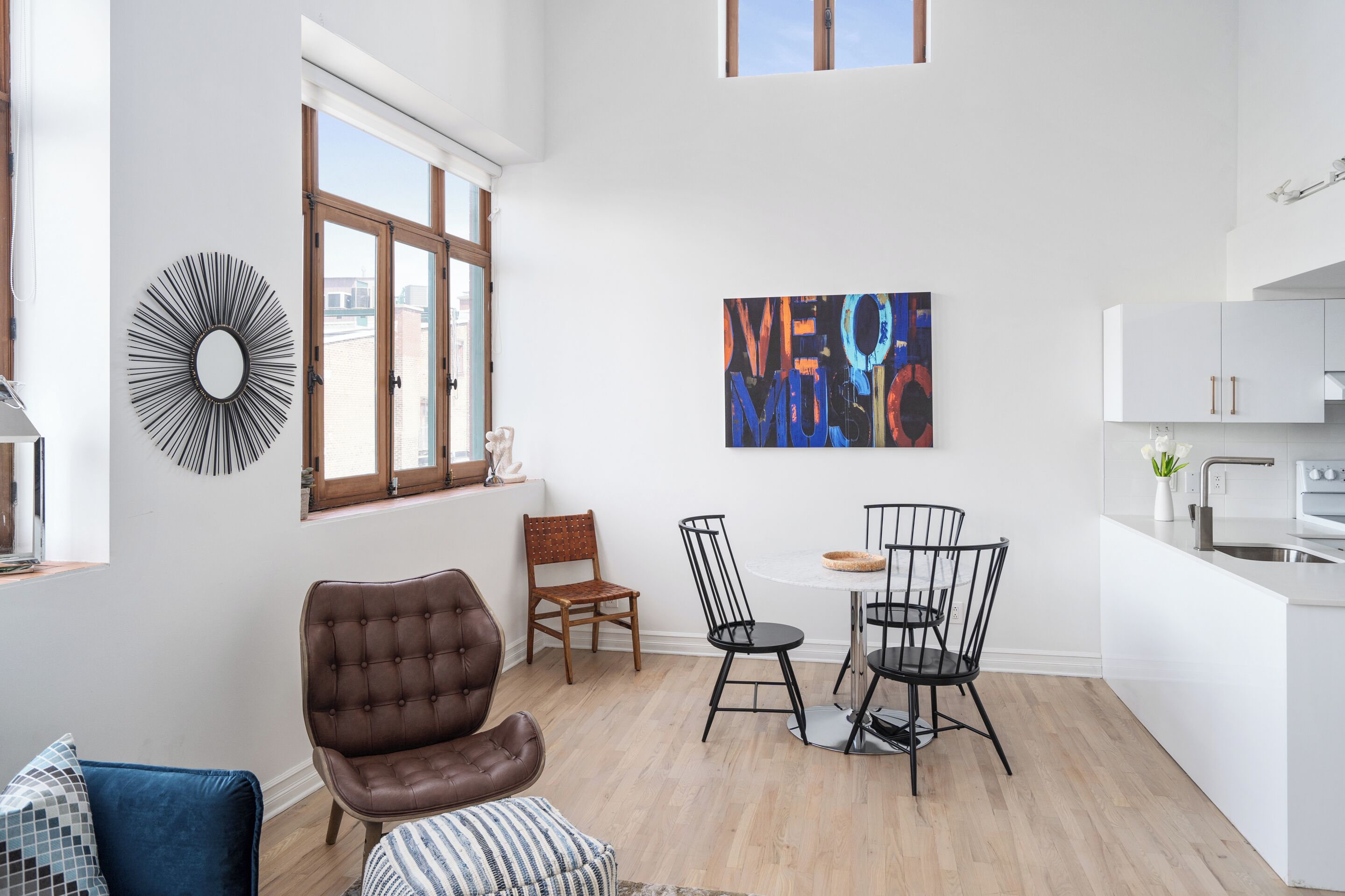 A modern kitchen with white cabinets, a dining area with a round table and chairs, and a large window with a view of buildings.