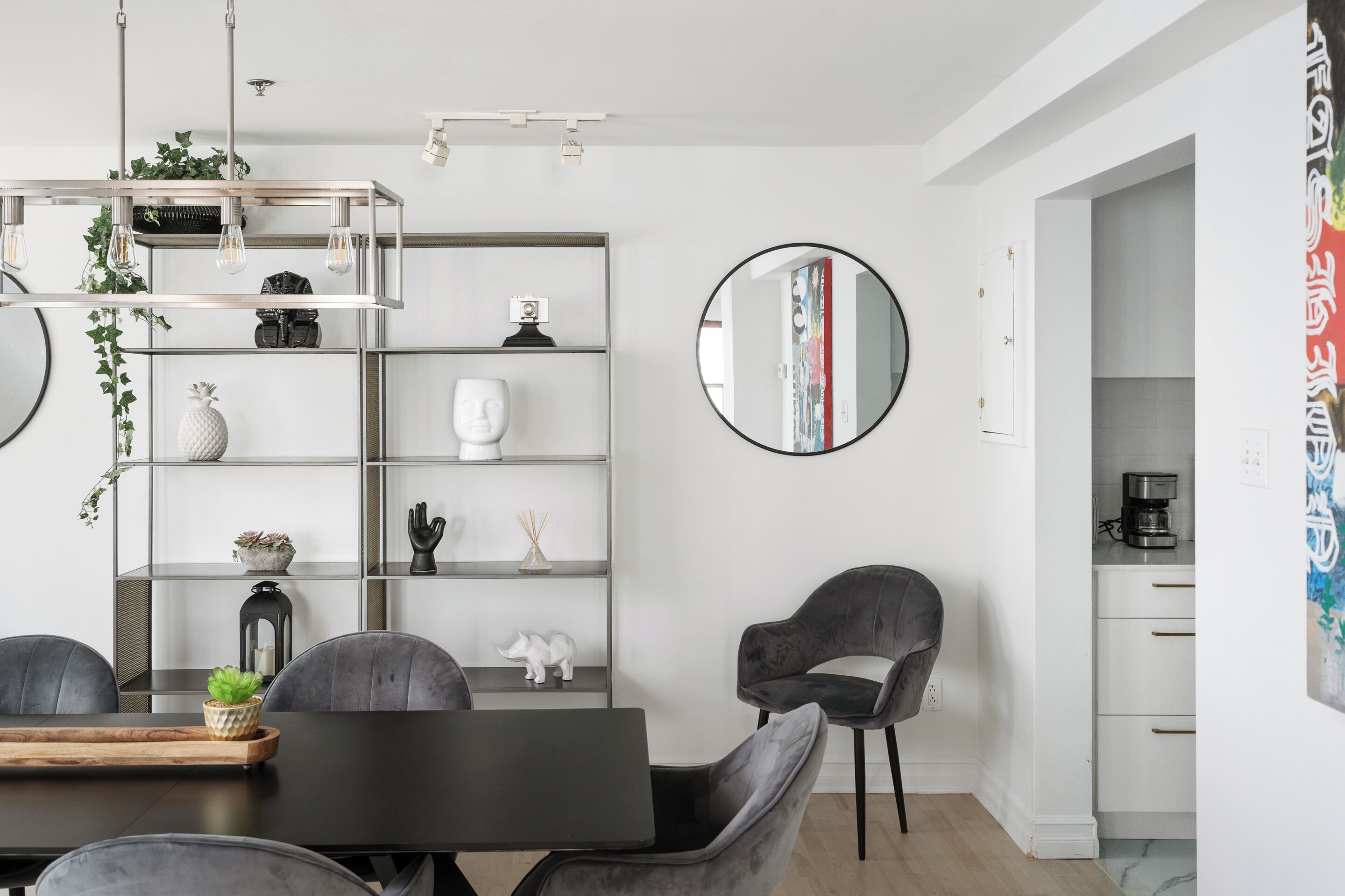 A modern dining area with a dark wooden table, grey upholstered chairs, a metal shelving unit with decorative items, and a round mirror on the wall.
