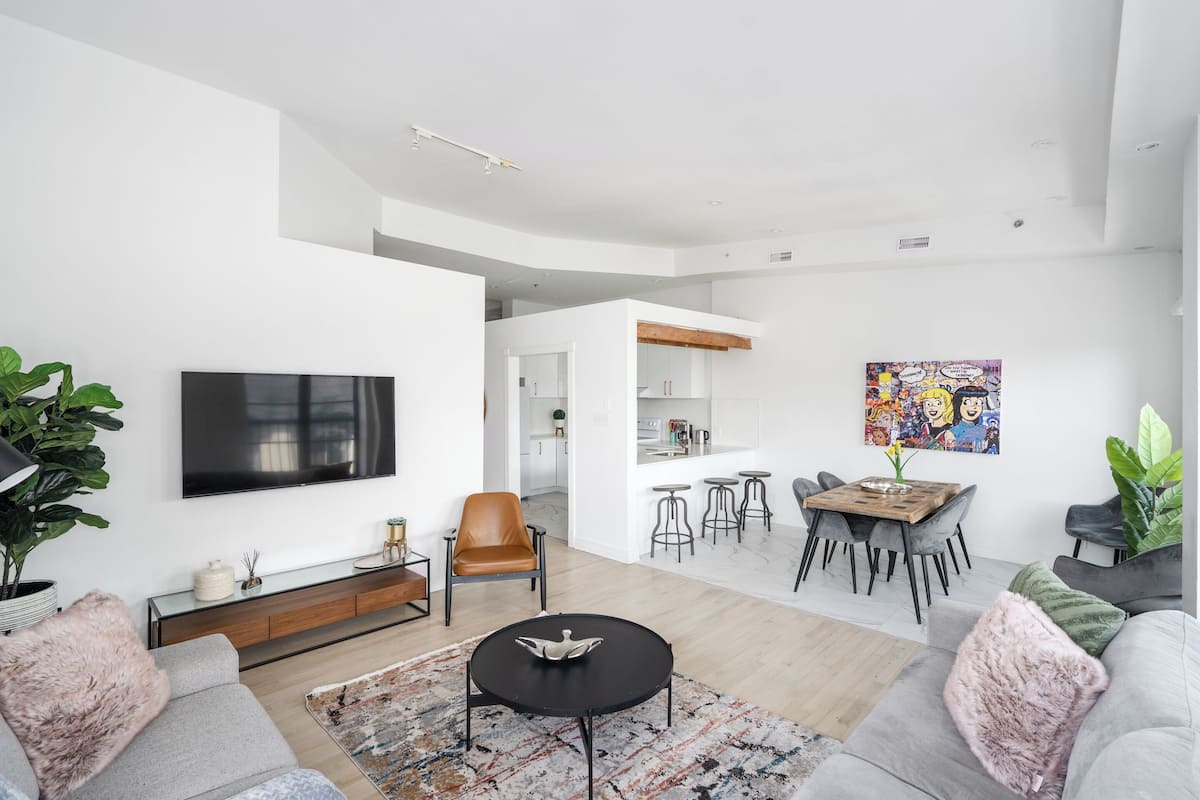 A modern living room with a flat-screen TV, a dining area with stools, and a kitchen in the background.