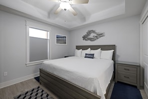 A bedroom with a bed, a nightstand, a wall-mounted artwork, a ceiling fan, and a window with blinds.