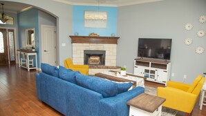 Ein Wohnzimmer mit einem blauen Sofa, gelben Sesseln, einem Kamin, einem Fernseher und einer Küchenzeile im Hintergrund.