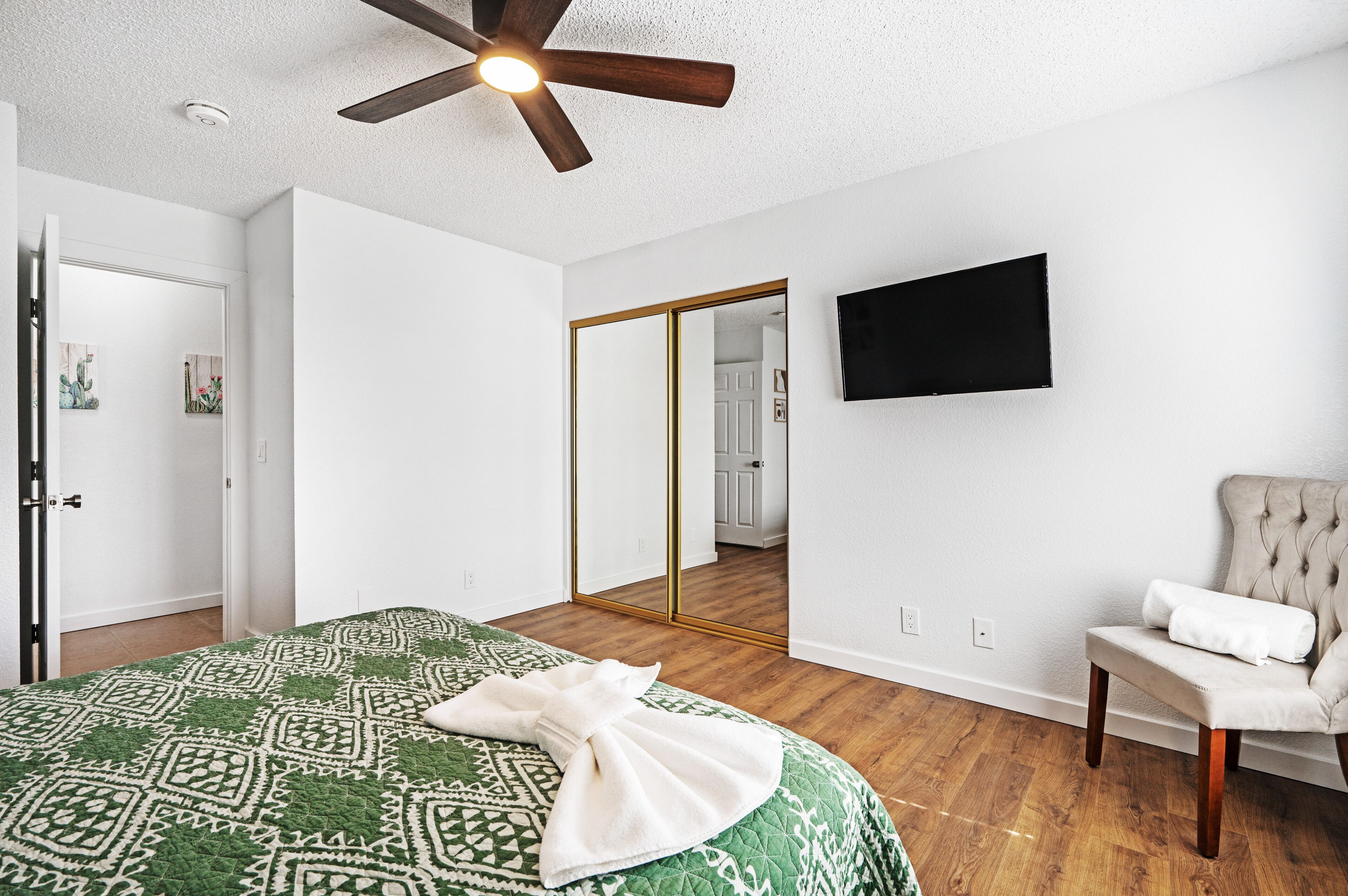 Une chambre à coucher avec un lit, un ventilateur de plafond, un miroir, une chaise et un téléviseur fixé au mur.