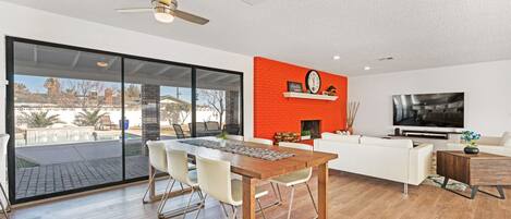 A modern living room with a dining area, a red accent wall, a large TV, and a view of a swimming pool.