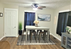 A dining area with a white table, four chairs, a ceiling fan, and a painting on the wall.