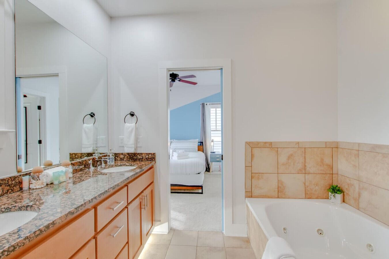 A modern bathroom with a large mirror, two sinks, a bathtub, and a visible bedroom with a bed and curtains.