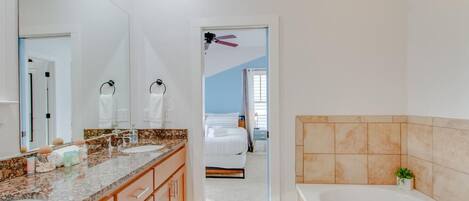 A modern bathroom with a large mirror, two sinks, a bathtub, and a visible bedroom with a bed and curtains.