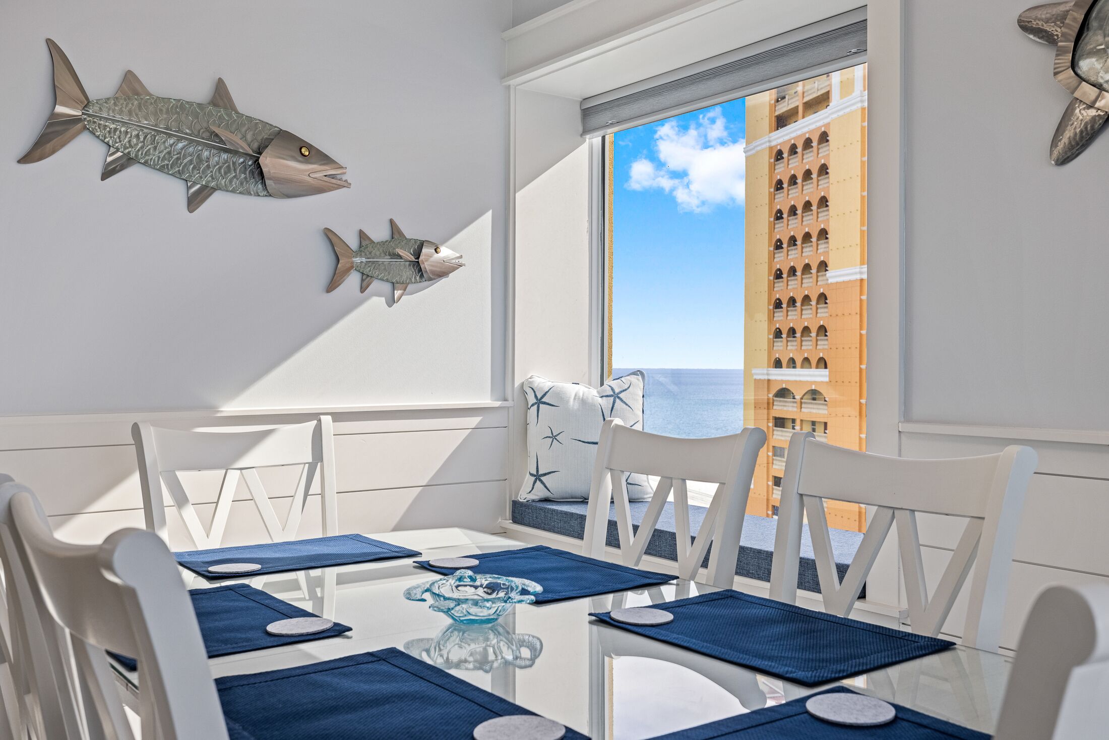 Un comedor con sillas blancas, manteles individuales azules y una mesa de vidrio. Hay dos adornos de pescado en la pared.