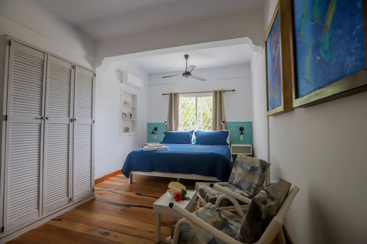 Un dormitorio con una cama, una silla, una mesita con una flor, un ventilador de techo y una ventana con cortinas.