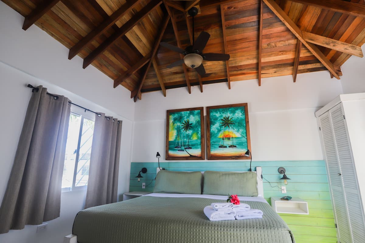 Un dormitorio con techo de madera, una cama con ropa de cama verde, dos cuadros en la pared y una ventana con cortinas.