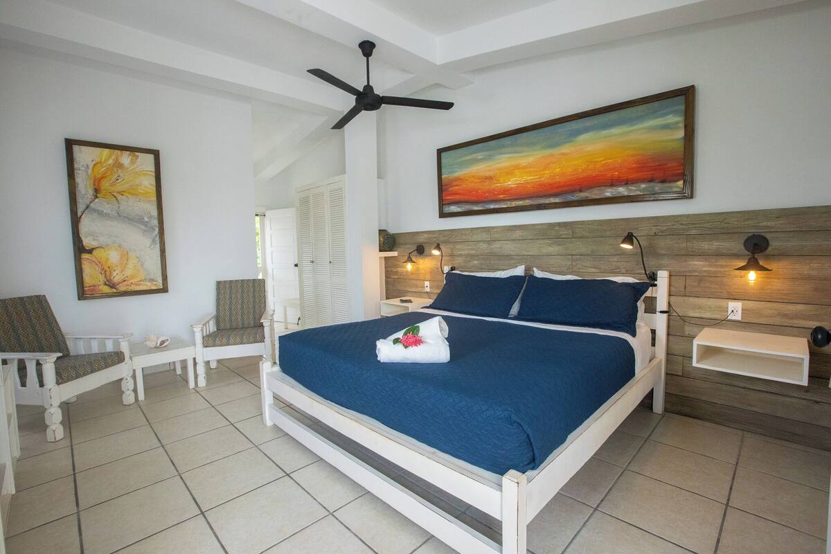 Un dormitorio con cama, ventilador de techo, un cuadro, una silla y una mesita de noche.