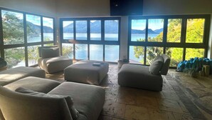 A spacious room with large windows offering a view of a lake and mountains. There are several sofas and a television mounted on the wall. - Lakeview on the Rocks (San Antonio Palopó)