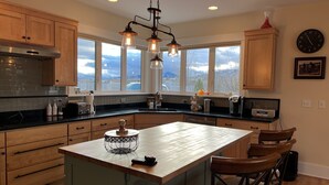 A modern kitchen with wooden cabinets, a large island, bar stools, and a view of the mountains through the windows. - Modern Dog Friendly Paradise Valley Home near Yellowstone NP & Chico Hot Springs (Pray)