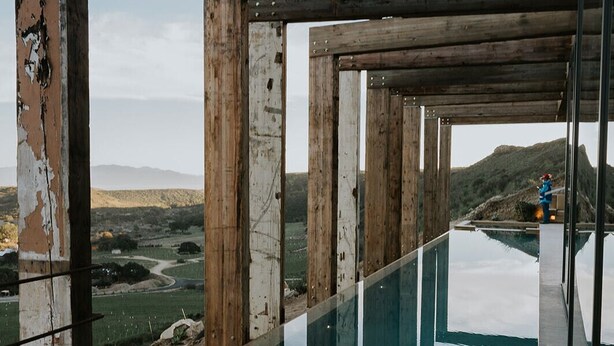 A modern pool with a clear glass bottom, surrounded by wooden beams and a view of a hilly landscape. - Vineyard Desert House -CuatroCuatros-CasaUno (Ensenada)
