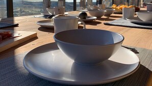 A dining table with bowls, cups, and a plate set for a meal, overlooking a scenic landscape through large windows. - Vineyard Desert House -CuatroCuatros-CasaUno (Ensenada)