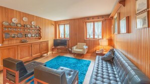 Une pièce aux murs lambrissés, avec un canapé, des fauteuils, un téléviseur et un tapis bleu.
